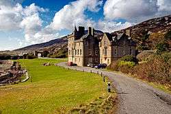 A large mansion house in the Scottish baronial style of the 19th century sits at the end of a narrow driveway with the shore on the left and moorland to the right.