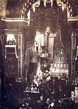 Looking down upon an assembly in a large, vaulted cathedral with a figure sitting on a large, canopied throne to the left of an altar