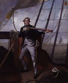 A large amn in a blue uniform strikes a dramatic pose holding a telescope on the quarterdeck of a ship. In the distance another ship can be seen with its sails set.