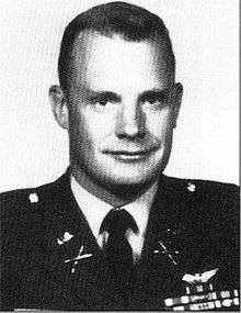 Head and shoulders of a white man with short hair and a slight smile, wearing a military jacket with ribbon bars and a winged pin on the left breast.