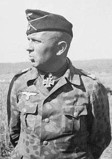 The head and shoulders of an elderly man, shown in partial profile. He wears a field cap and a military uniform and an Iron Cross displayed at the front of his camouflage shirt collar.