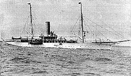 A black and white image of a twin-masted motor yacht with a funnel afloat with no sails set.