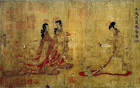 Two ladies walking towards another lady standing at a writing table with a writing brush in her hand