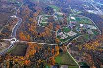An aerial view of a school campus in a wooded area. The trees around the campus have red leaves. Roads cut through the middle of the wood toward the school.
