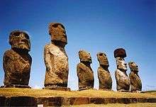 Row of six large stone statues with elongated heads on a grassy slope