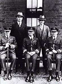 Formal portrait of five men, three seated and wearing military uniforms with peaked caps, two standing and wearing civilian clothes and hats