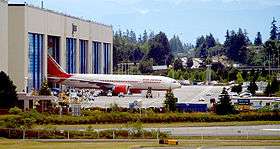 Aircraft factory. Rectangular building with six doors, one of which is open to reveal an emerging airliner. The background is a forest; small vehicles and taxiways surround the factory.