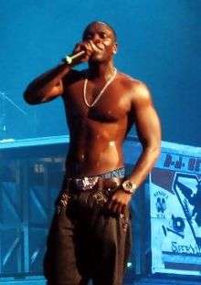 Akon shirtless wearing black pants, singing into a microphone on stage.