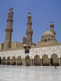 A paved courtyard is visible in the foreground, and behind it a wall of angular keel-shaped arched bays supported by columns. Behind the wall, two minarets, a dome, and another minaret are visible from left to right. In the far background in the center the top of another minaret can be seen.