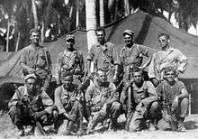 Black-and-white image of ten men in two rows, the top row standing and the bottom row crouching, are all facing the camera. They are wearing military attire and are holding rifles.