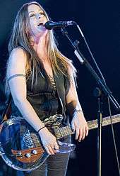 A Caucasian female with light colored hair leaning her head back while singing into a microphone. She is wearing a sleeveless green shirt with dark grey pants and has a guitar strapped around her.