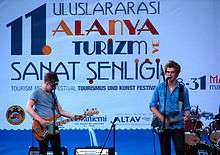 Two guitarists in blue shirts perform on stage in front of a blue and white poster in Turkish.