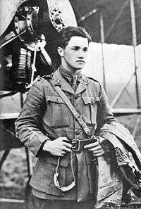Half portrait of young dark-haired man in military uniform with coat over left arm, standing in front of biplane