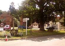 Photograph of a small village green, showing a drinking fountain, oak tree, and well