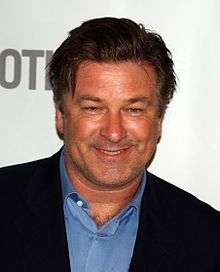 Alec Baldwin, a caucasian male in his early-50s with dark uncombed hair, wears a black suit with a blue shirt with the collar open. He smiles and stands in front of a white background with grey font.