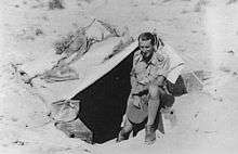 Man in shorts and long socks holding his slouch hat in his hand, in front of a small tent stretched over a hole in the sand.