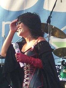 A dark-haired woman, wearing a black corset with red applications and a black cloak, holding a microphone in her left hand and touching her face with her right hand.