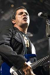 A man with a black jacket, black shirt with gold accents, singing in a microphone with his hand on a blue guitar.