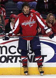 An unshaven man with black hair stares into the distance. He is wearing a red uniform with blue pants and a logo saying "Washington Capitals" on his chest.
