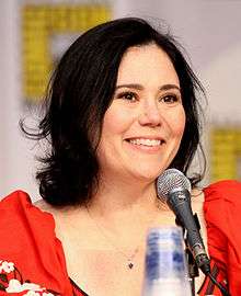 A caucasian woman with black hair tied back, smiling into a microphone, with a vague symbol behind her.