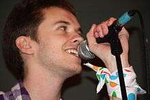A close-up photograph of Alex Day singing into a microphone, taken from below.