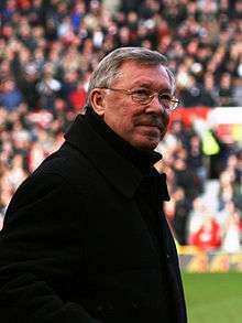 The torso and head of a grey-haired white man in a football stadium. He is wearing spectacles and a black coat.