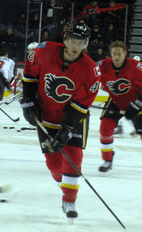 A hockey player in a red uniform with black trim and a stylized black "C" logo on the chest skates toward the goal