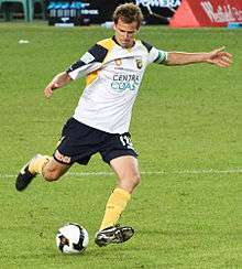 A man wearing a white shirt and navy shorts in the act of kicking a football with his right foot.