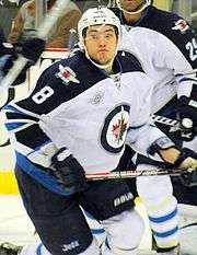 A Caucasian hockey player. He is wearing a white jersey with blue shoulders with a jet over a maple leaf inside of a circle for the logo. He is starting his skating stride with his eyes looking towards the camera.