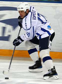An ice hockey player skating on the ice while using his hockey stick to control the puck. He is wearing a white helmet and a white and black uniform.