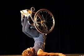 A woman in dark, close-fitting clothes and a wheelchair lies on her back, stabilizing herself with her arms and lifting her legs, along with the wheelchair, over her head