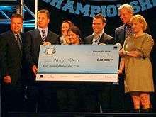 A high school student stands behind a large blue check. Behind her stand six adults in suits. The check has a National Vocabulary Championship logo in the top left and is written out to Aliya Deri for the amount of $40,000. The check is dated March 10, 2008.