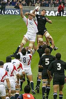 The All Blacks and England contesting a line-out. Both sets of forwards lined up wearing white and black respectively, with a player from each side at the rear of the line out being lifted by their team-mates while both reaching for the ball.
