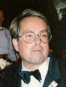 The photograph of a bespectacled man wearing a tuxedo with a white and black pocket square in his left chest pocket.