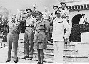 Eight men standing on the steps of a Middle Eastern building. Six are wearing various uniforms but one is wearing a business suit.