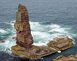 A tall sea stack stands on a wave-lashed rocky platform of rocks.