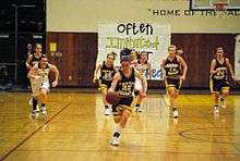 A female high school basketball player dribbles the ball towards the camera with a focused look on her face. Behind her trail basketball members from her team and the opposing team. All are running towards the camera.