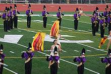 Two color guard members dressed in togas spin flags with fire designs in the middle of the DVC football field. Surrounding these color guard members are flute and saxophone players standing still and playing. They are dressed in a purple jacket and black marching pants and are wearing shakos.