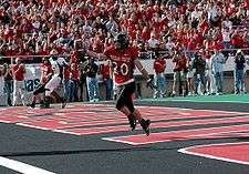 Texas Tech Red Raider football player stands with the a football in the endzone in the foreground with fans in the stands in the background cheering.