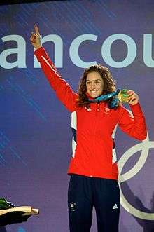 A brunette with long wavy hair smiles at the top of a podium as she raises her right arm and holds, with her left hand, a gold medal hung around her neck. She wears a vivid red sports jacket and dark blue sports trousers.