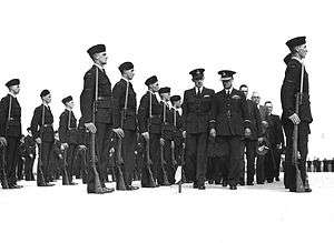 Two columns of men, some in military uniforms with peaked caps, some in civilian clothes, walking between two rows of troops with forage caps standing to attention with rifles and fixed bayonets, and further rows of troops behind them