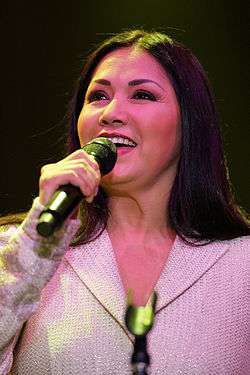 A woman with black hair, facing front, holding a microphone in her left hand, behind a microphone pedestal.
