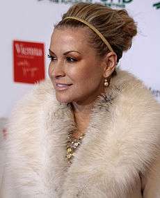 A woman in makeup smiles. Her locks are held back by a hairband, and she wears an elaborate fur-like material arouns her neck.