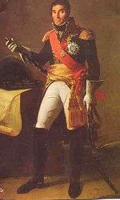Formal full-length portrait of Masséna in dress military uniform, comprising white breeches with knee length black boots, dark cutaway coat with high collar and gold embroidery, a red shoulder sash and gold waist sash. He wears a large star of honor on his breast. He is a tall dark man with a long face and thick eyebrows. He looks quizzically at the observer and holds a marshal's baton, and saber.