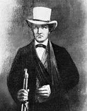 A man in a dark suit with a white hat. His left arm seems to be in a sling. In his right hand he grasps the barrel of a rifle.