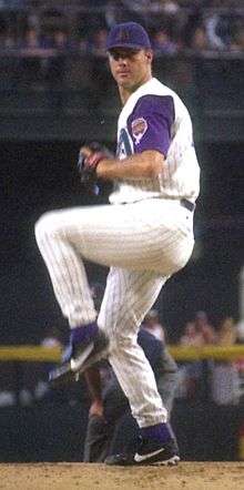 A man in a white pinstriped baseball uniform with purple sleeves and cap stands with his left leg lifted as if about to pitch a baseball.