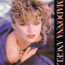 Madonna with rough, scrambled blond hair. She wraps a blue cloth around herself with her hands closing it on her chest.