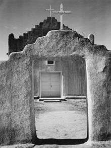 A black-and-white vertical photograph shows an adobe wall in the foreground, rising in the middle with a stair-step pattern and a white wooden cross at the pinnacle, with an open doorway beneath. Through the doorway and above the wall, an adobe church with white double doors and a similar stair-stepped roof and cross stands, slightly larger than the wall in front of it. The midday sun casts harsh shadows on the dirt ground.