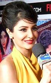 Anushka Sharma holding a mike and smiling away from the camera. She is dressed in a halter-neck yellow outfit.