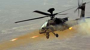 Colour photograph of an Apache Attack helicopter at right of image facing left, firing two rockets at a target beyond the bottom left of the frame.